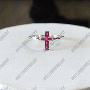 May include: A silver ring with a pink cross made of small rectangular stones. The ring is on a white surface.