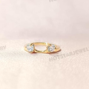 Op de afbeelding: Een goudkleurige ring met twee heldere, ronde edelstenen. De ring heeft een uniek ontwerp, met de edelstenen aan weerszijden van een open ruimte. De ring wordt gepresenteerd op een lichtroze oppervlak.