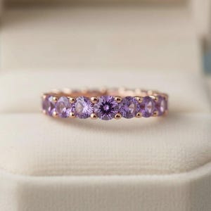 May include: A rose gold ring featuring a series of oval-cut, light purple gemstones. The gemstones are set closely together, creating a continuous band of color. The ring is displayed on a soft, white surface.