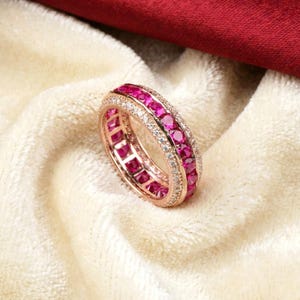 May include: Rose gold ring with a double row of vibrant pink gemstones and a single row of smaller, clear gemstones. The ring is set against a soft, off-white fabric with a deep red fabric in the background. The ring is a piece of jewelry.
