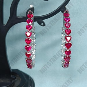 以下が含まれることがあります： 赤いハート型の石と透明なハート型の石が並んだ、銀製のフープイヤリングのペアです。