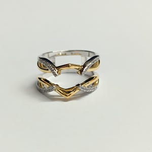 May include: A two-tone ring guard featuring a chevron-shaped design with polished yellow gold bands and silver-toned accents encrusted with small clear stones. The ring has an open center gap and a split shank construction with intertwined metal details.