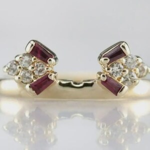 May include: A gold-colored ring with a unique design. The ring features two clusters of gemstones, each with a mix of small, round, clear stones and rectangular, deep red stones. The ring is set against a soft, reflective background.