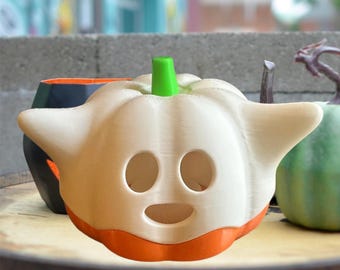 May include: A white ceramic pumpkin with a green stem and orange base. The pumpkin has a ghost face with two eyes and a mouth.