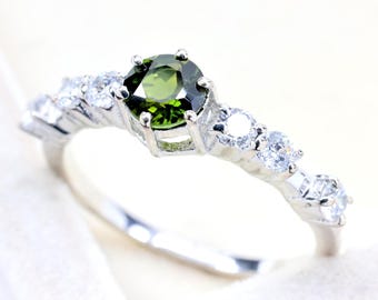 May include: A silver-toned ring featuring a round, faceted olive-green center stone held by six prongs. The band is accented with three clear, round, faceted stones on each side, creating a symmetrical design with a polished metallic finish.