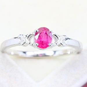 May include: A silver-toned ring featuring a central oval-cut vibrant pink gemstone held by four prongs. The band displays a decorative, textured design on both sides of the stone, transitioning into a smooth, polished metal shank.