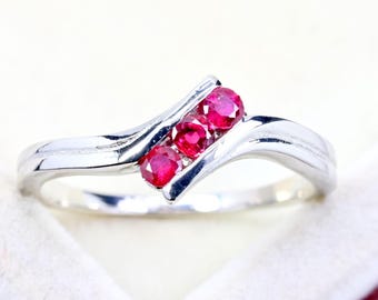 May include: A silver-toned ring featuring a bypass-style band with a polished finish. The center of the ring is set with three round, vibrant red rubies arranged in a diagonal row, held securely within the metal setting of the band.