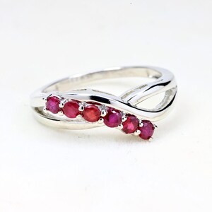 May include: A silver-toned ring featuring a bypass-style band with a polished finish. The ring is set with six small, round, vibrant red rubies arranged in a single row along one side of the overlapping band, creating a sleek and continuous design.