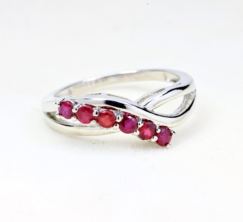 May include: A silver-toned ring featuring a bypass-style band with a polished finish. The ring is set with six small, round, vibrant red rubies arranged in a single row along one side of the overlapping band, creating a sleek and continuous design.