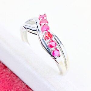 May include: A silver-toned ring featuring a crossover band design with five round, vibrant pinkish-red gemstones set in a vertical row along the center. The polished metal band curves gracefully, creating an intertwined appearance around the stones.