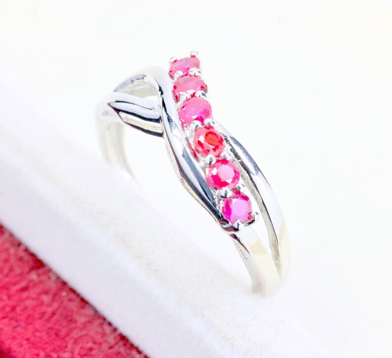 May include: A silver-toned ring featuring a crossover band design with five round, vibrant pinkish-red gemstones set in a vertical row along the center. The polished metal band curves gracefully, creating an intertwined appearance around the stones.