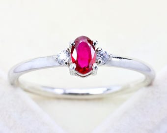 May include: A silver-toned ring featuring a central oval-cut vibrant red gemstone held in a prong setting, flanked by two small clear round-cut stones on either side of the band. The metal band is smooth, polished, and has a reflective finish.