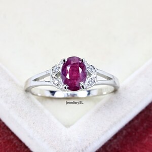 May include: A silver ring featuring a red oval ruby gemstone surrounded by smaller white gemstones. The ring is displayed against a white backdrop.