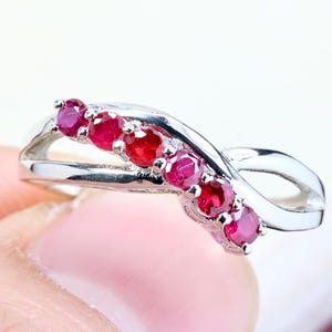 May include: A silver-toned ring featuring a crossover band design set with five round, deep red rubies arranged in a curved line along the center. The metal band has a polished, reflective finish and a smooth, flowing shape that tapers at the ends.