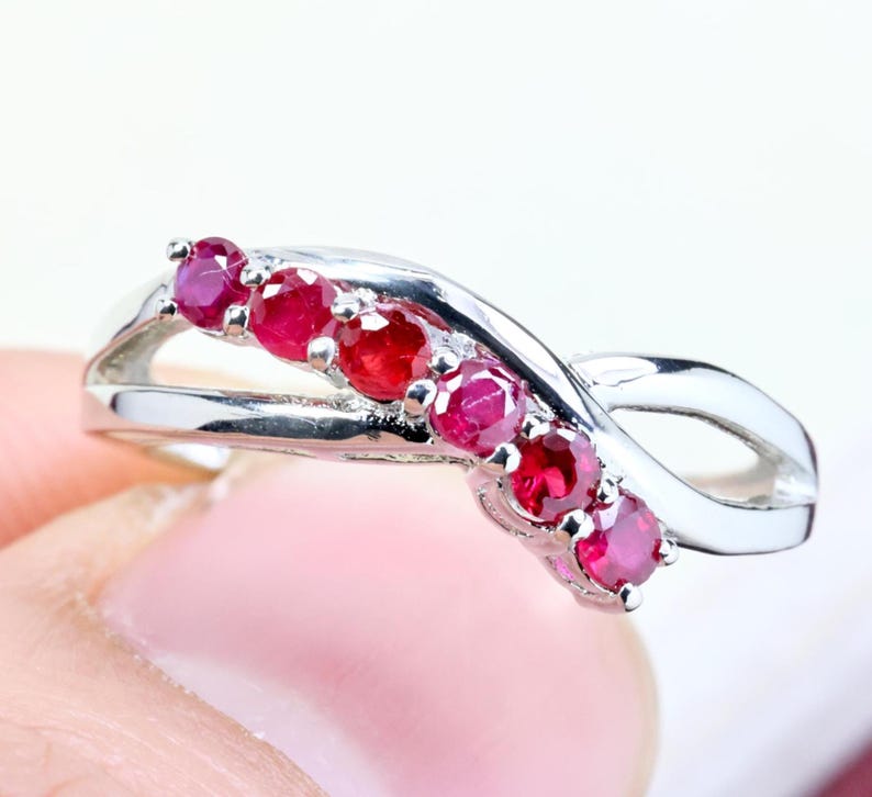 May include: A silver-toned ring featuring a crossover band design set with five round, deep red rubies arranged in a curved line along the center. The metal band has a polished, reflective finish and a smooth, flowing shape that tapers at the ends.