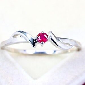 Puede incluir: Un anillo de tono plateado que presenta una única piedra preciosa de rubí rojo vibrante, de corte redondo, engastada en el centro. La banda tiene un acabado liso y pulido con un diseño curvo en forma de onda que se estrecha hacia el engaste.