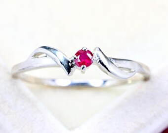 May include: A silver-toned ring featuring a single, small, round-cut vibrant red ruby gemstone set in the center. The band has a smooth, polished finish with a curved, wave-like design that tapers toward the central stone setting.