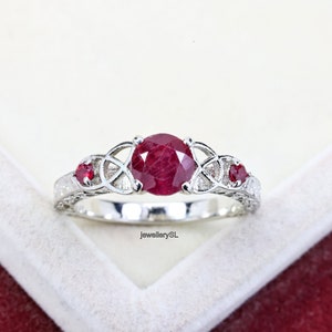 May include: A silver ring featuring a large red ruby gemstone and two smaller red ruby gemstones on either side. The ring has a Celtic knot design.