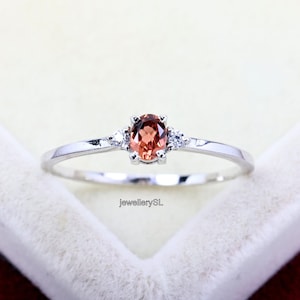 May include: A silver ring featuring a small oval-shaped orange gemstone, flanked by two smaller white gemstones. The ring is displayed on a white surface.