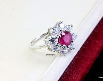 May include: A silver ring featuring a large red ruby gemstone surrounded by smaller white gemstones. The ring is set in a flower-shaped design.