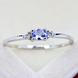 May include: A silver-toned ring featuring a central oval-cut light purple gemstone flanked by two small, clear, round-cut accent stones. The band is thin and polished, showcasing a delicate design with a prong setting for each of the three stones.