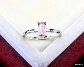 May include: A silver-toned ring featuring a single emerald-cut light pink gemstone set in a four-prong setting. The band is smooth and polished, showcasing a minimalist design with the text JewellerySL visible near the base of the stone.