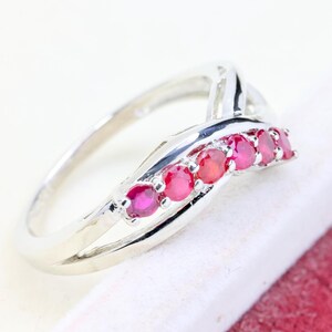 May include: A silver-toned ring featuring a crossover band design with five round, vibrant red rubies set in a row along the top curve. The metal band has a polished, reflective finish and a smooth, continuous structure that tapers toward the back.