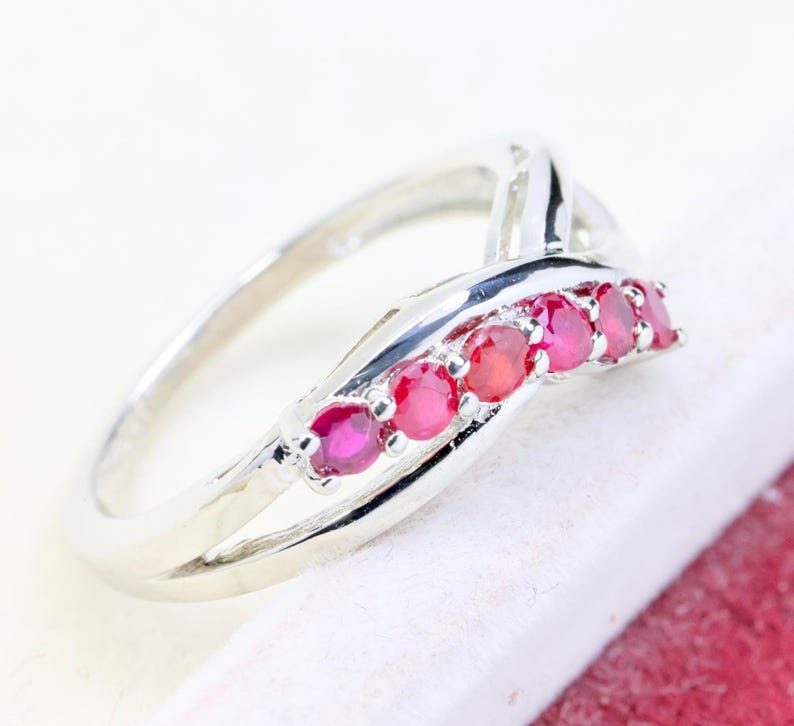 May include: A silver-toned ring featuring a crossover band design with five round, vibrant red rubies set in a row along the top curve. The metal band has a polished, reflective finish and a smooth, continuous structure that tapers toward the back.