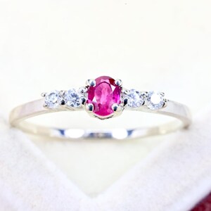 May include: A silver-toned ring featuring a central oval-cut vibrant pinkish-red gemstone held by four prongs, flanked on each side by two small, round, clear white stones set into the polished metal band.