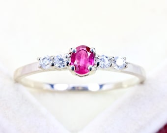 May include: A silver-toned ring featuring a central oval-cut vibrant pinkish-red gemstone held by four prongs, flanked on each side by two small, round, clear white stones set into the polished metal band.