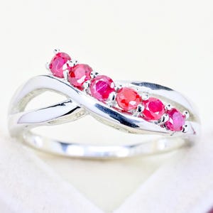 May include: A silver-toned ring featuring a crossover band design with five round, vibrant pinkish-red gemstones set in a row along the top curve. The metal band has a polished, reflective finish that creates a smooth, continuous loop structure.