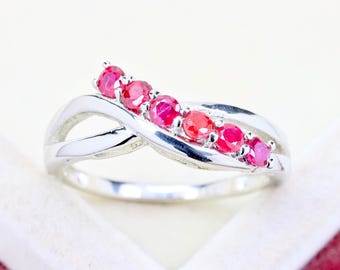May include: A silver-toned ring featuring a crossover band design with five round, vibrant pinkish-red gemstones set in a row along the top curve. The metal band has a polished, reflective finish that creates a smooth, continuous loop structure.