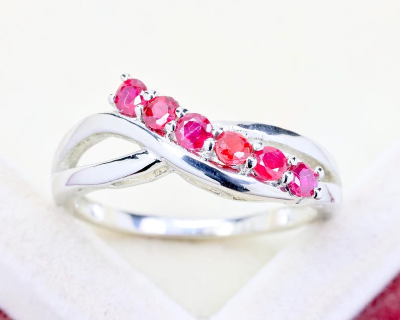 May include: A silver-toned ring featuring a crossover band design with five round, vibrant pinkish-red gemstones set in a row along the top curve. The metal band has a polished, reflective finish that creates a smooth, continuous loop structure.