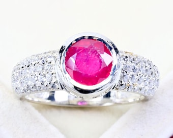 May include: A silver-toned ring featuring a large, round, faceted vibrant red gemstone set in a smooth bezel. The band is wide and encrusted with numerous small, clear, sparkling stones that extend across the shoulders of the ring.