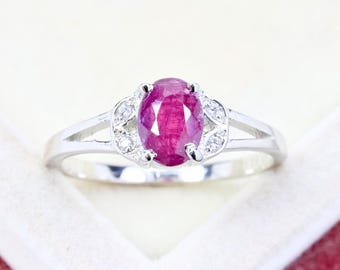 Puede incluir: Un anillo de tono plateado con una piedra preciosa central de corte ovalado en color rojo rosado intenso, sujeta por cuatro garras. La banda se divide en dos brazos a cada lado, decorados con dos pequeñas piedras claras redondas en diseño de hoja.