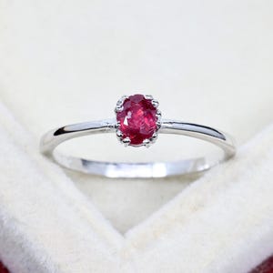 Puede incluir: Un anillo fino de tono plateado con una piedra preciosa de color rojo intenso, de corte ovalado y engastada en garras. La banda es lisa y pulida, mostrando un diseño sencillo que resalta el color vibrante y la textura facetada de la piedra.