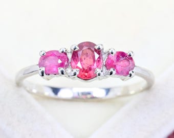 Pode incluir: Um anel de prata com três pedras preciosas de cor rosa vibrante em corte oval dispostas numa fila horizontal. A pedra central é ligeiramente maior do que as duas laterais, cada uma fixada por quatro garras numa banda de prata polida.