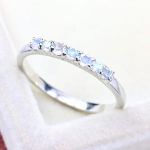 Op de afbeelding: Een zilverkleurige ring met een rij van zeven kleine, ronde, iriserende maanstenen in een klauwzetting. Het metaal heeft een gepolijste, reflecterende afwerking en de stenen vertonen een zachte, doorschijnende blauwe gloed over het oppervlak.