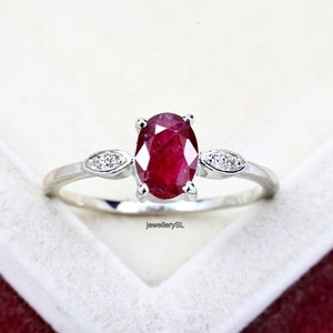 Puede incluir: Un anillo de plata con una gran piedra de rubí de forma ovalada y dos pequeños acentos de diamantes a cada lado. El anillo está colocado sobre un fondo blanco.