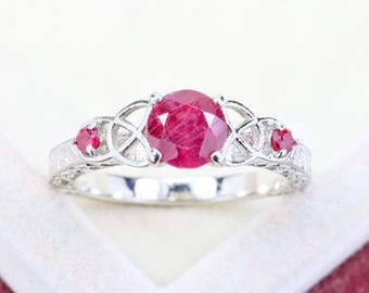 May include: A silver ring featuring a large, round, faceted red ruby center stone flanked by two smaller round red rubies. The band is crafted with intricate openwork metal detailing that creates a decorative pattern around the stones.