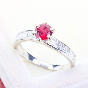 Könnte beinhalten: Ein silberfarbener Ring mit einem runden, facettierten roten Mittelstein in einer Sechser-Krappenfassung. Die Ringschiene ist mit einer Reihe kleiner, klarer, quadratisch geschliffener Steine besetzt, die beidseitig des roten Edelsteins liegen.