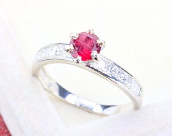 May include: A silver-toned ring featuring a round, faceted red centre stone held by a six-prong setting. The band is accented with a channel of small, clear, square-cut stones embedded along the shank on both sides of the central red gemstone.
