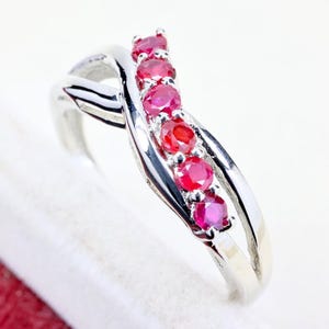 May include: A silver-toned ring featuring a crossover band design with a row of six round, vibrant red rubies set in prongs along the center. The polished metal band curves gracefully, creating an intertwined appearance that highlights the stones.