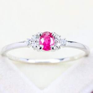 May include: A silver-toned ring featuring a central oval-cut vibrant pinkish-red gemstone. The center stone is flanked on each side by a cluster of three small, clear, round-cut accent stones set into the polished metal band.