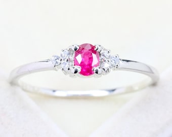 May include: A silver-toned ring featuring a central oval-cut vibrant pinkish-red gemstone. The center stone is flanked on each side by a cluster of three small, clear, round-cut accent stones set into the polished metal band.