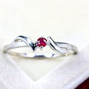 Peut inclure: Une bague en argent au design fluide, ornée d'une petite pierre précieuse rouge vif et ronde au centre. La bague a une finition polie et est posée sur un fond blanc doux. Il s'agit d'un bijou.