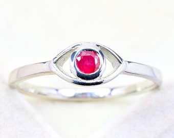 Könnte beinhalten: Ein silberner Ring mit einem zentralen, rund geschliffenen roten Rubin, der in einen augenförmigen Metallrahmen gefasst ist. Die Ringschiene ist glatt und poliert, wobei der Edelstein in einer Zargenfassung in der Mitte des ovalen Designs sitzt.