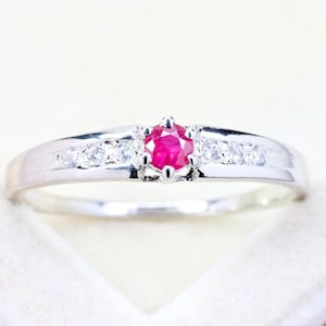 May include: A silver-toned ring featuring a central round-cut vibrant red ruby gemstone held in a six-prong setting, flanked by three small clear white stones on each side of the band, creating a symmetrical and polished appearance.