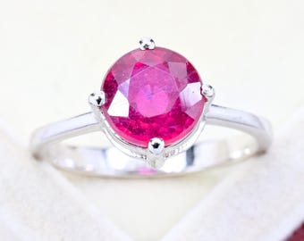 May include: A silver-toned ring featuring a round, faceted, vibrant pinkish-red gemstone set in a four-prong mounting. The band is smooth and polished, showcasing a clean, metallic finish that complements the central stone's bright, saturated colour.