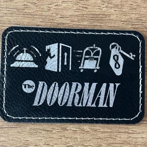 May include: A rectangular black patch with white stitched edges featuring a silver graphic print of a service bell, an open door, a luggage cart, and a hotel key tag with the number eight. The text "The DOORMAN" is printed in silver below.